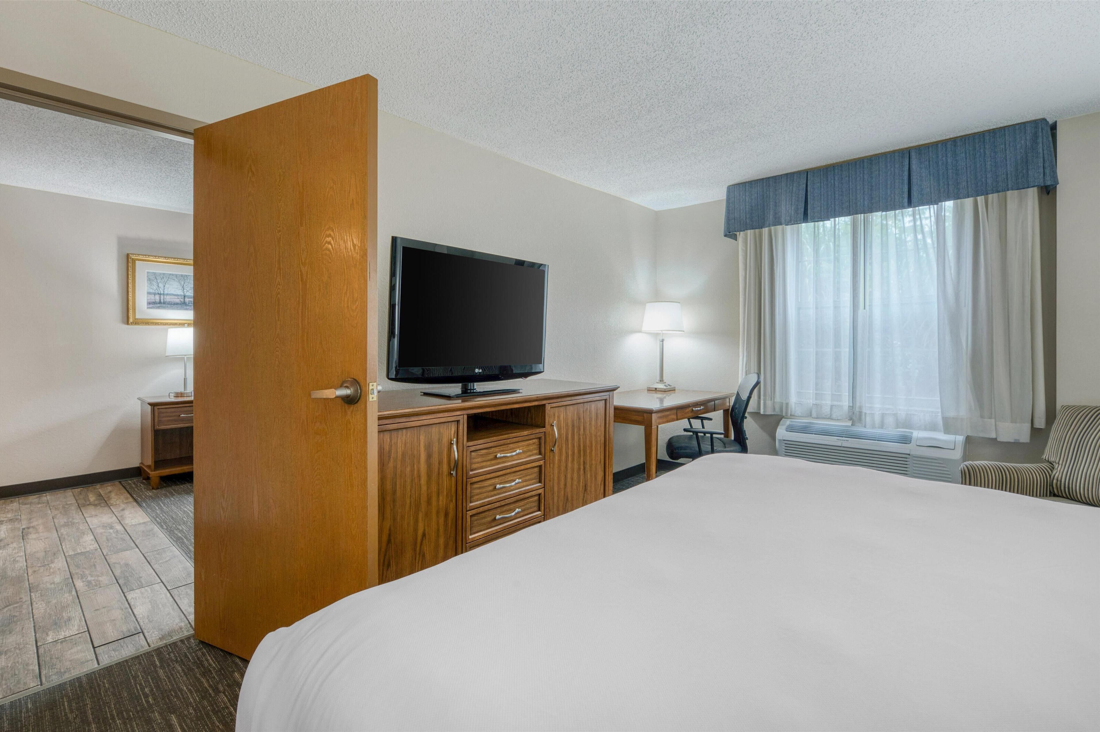 A hotel room with a large bed, a wooden desk with a flat-screen TV, a desk lamp, a chair, and a window with curtains.