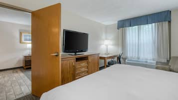 A hotel room with a large bed, a wooden desk with a flat-screen TV, a desk lamp, a chair, and a window with curtains.