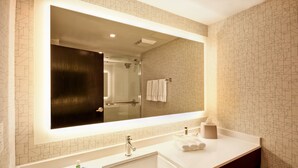 Une salle de bain avec un grand miroir, un lavabo blanc, une douchette à main et un porte-serviettes.