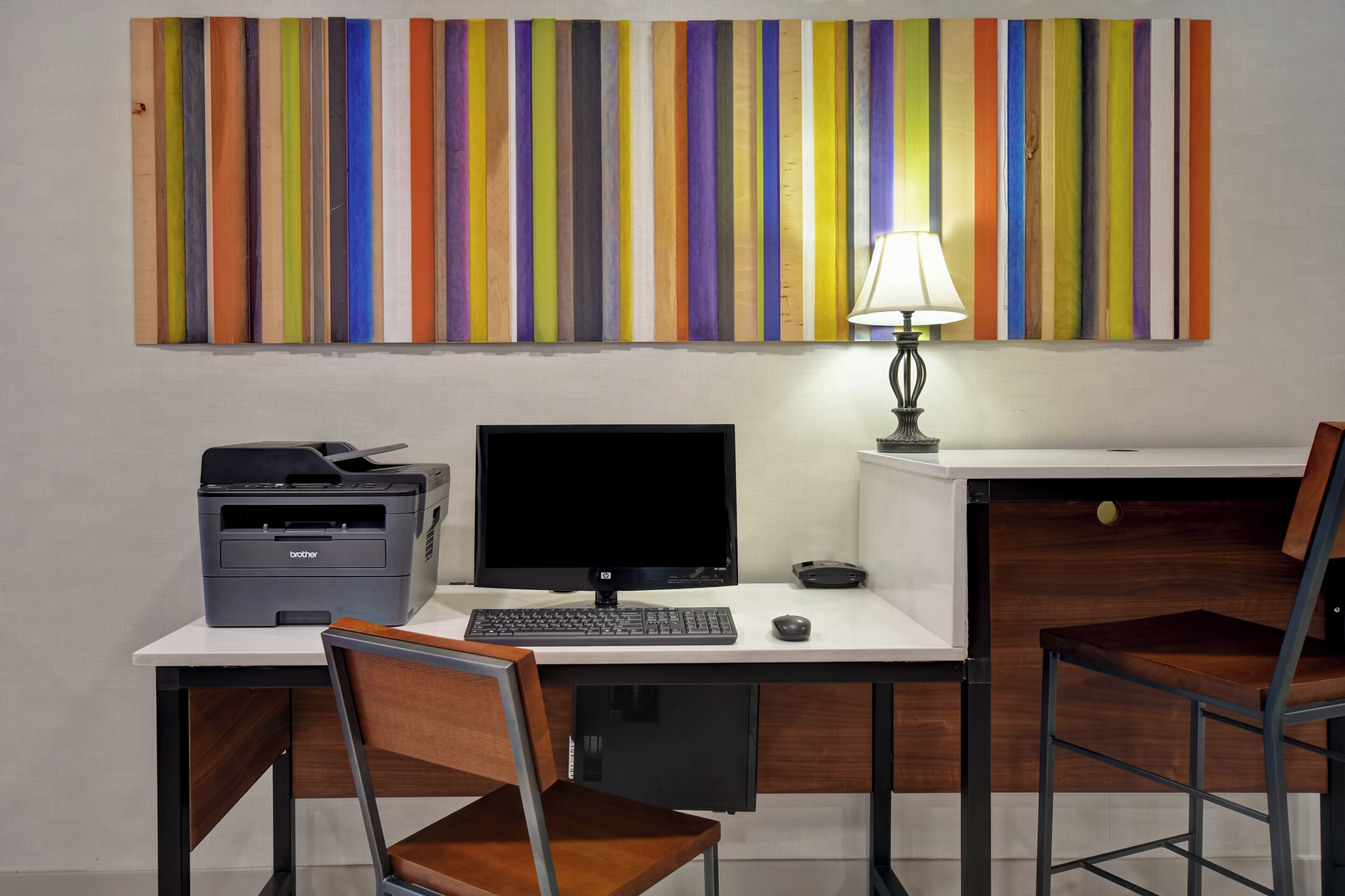 A workspace with a desk, computer, printer, and two chairs. A colorful striped artwork is on the wall above the desk.