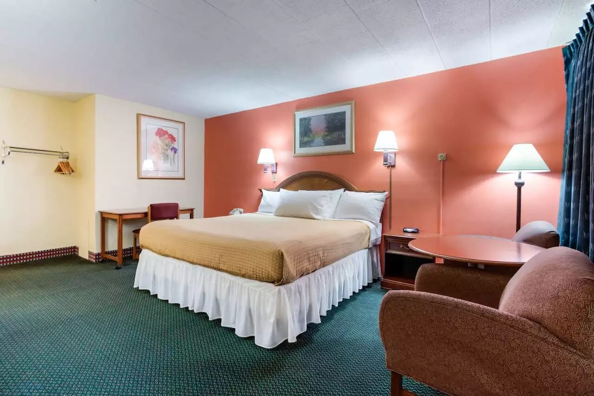 A hotel room with a bed, two bedside lamps, a small table, a chair, and a painting on the wall.