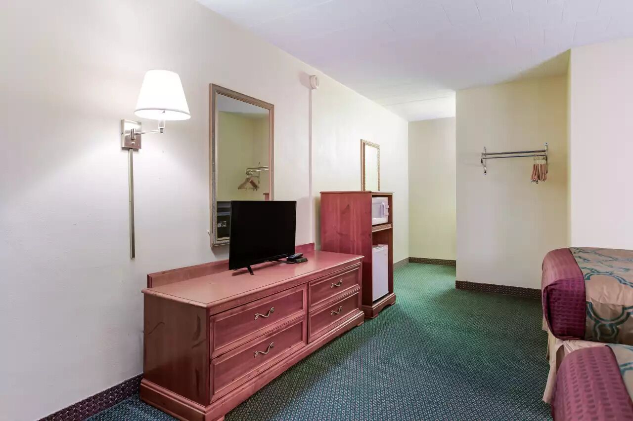 Quarto de hotel com cama, televisão sobre uma cômoda de madeira, espelho, luminária e cabideiro.