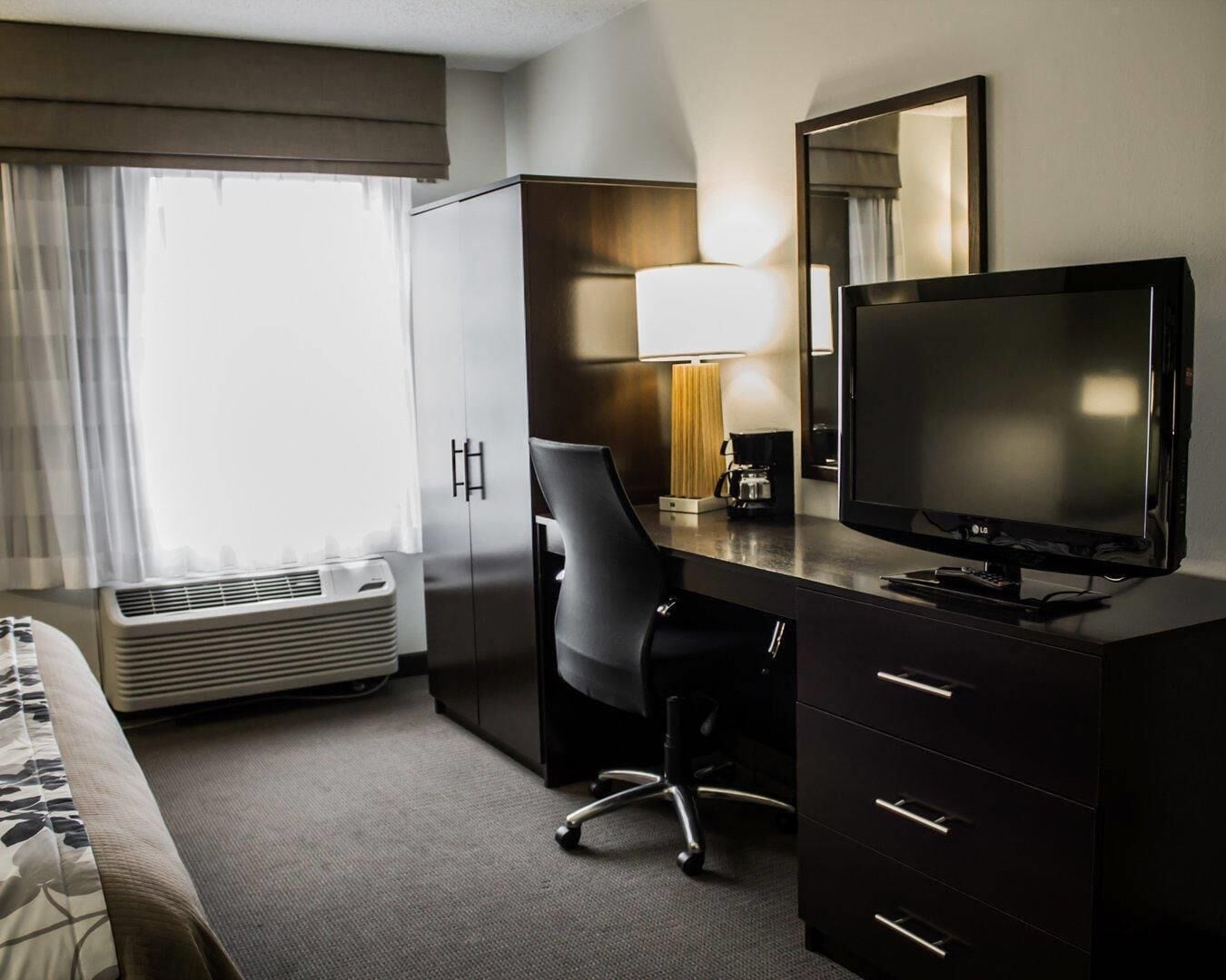 A hotel room with a bed, a desk with a TV, a chair, a lamp, and a wardrobe.