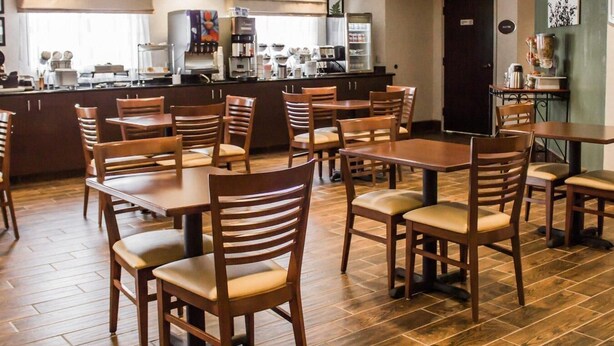 A restaurant with wooden tables and chairs, a kitchen area, and a green wall with a picture. - Suburban Studios Johnstown - Richland Town Center (Johnstown)