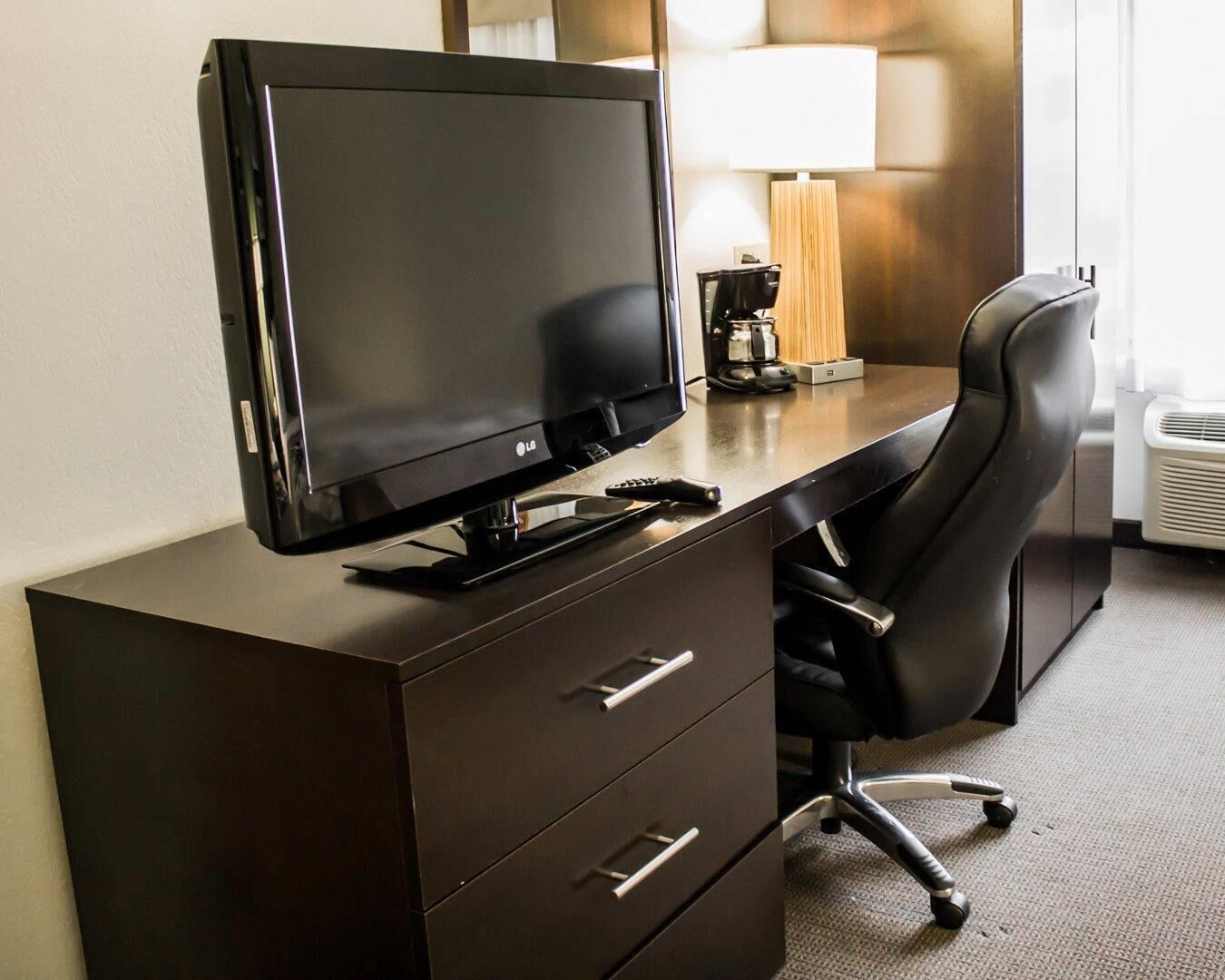 Habitación de hotel con escritorio, televisión de pantalla plana, silla, lámpara y cafetera.