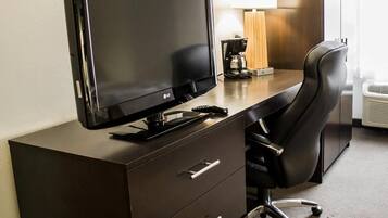 Habitación de hotel con escritorio, televisión de pantalla plana, silla, lámpara y cafetera.