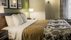 A hotel room with a bed, a nightstand, a lamp, a wall-mounted picture, and a window with curtains. - Suburban Studios Johnstown - Richland Town Center (Johnstown)