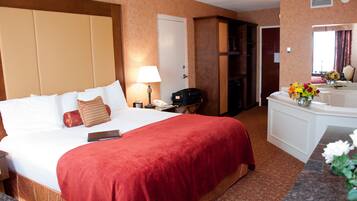 A hotel room with a large bed, a desk with a lamp, a minibar, and a bathroom with a mirror and flowers.
