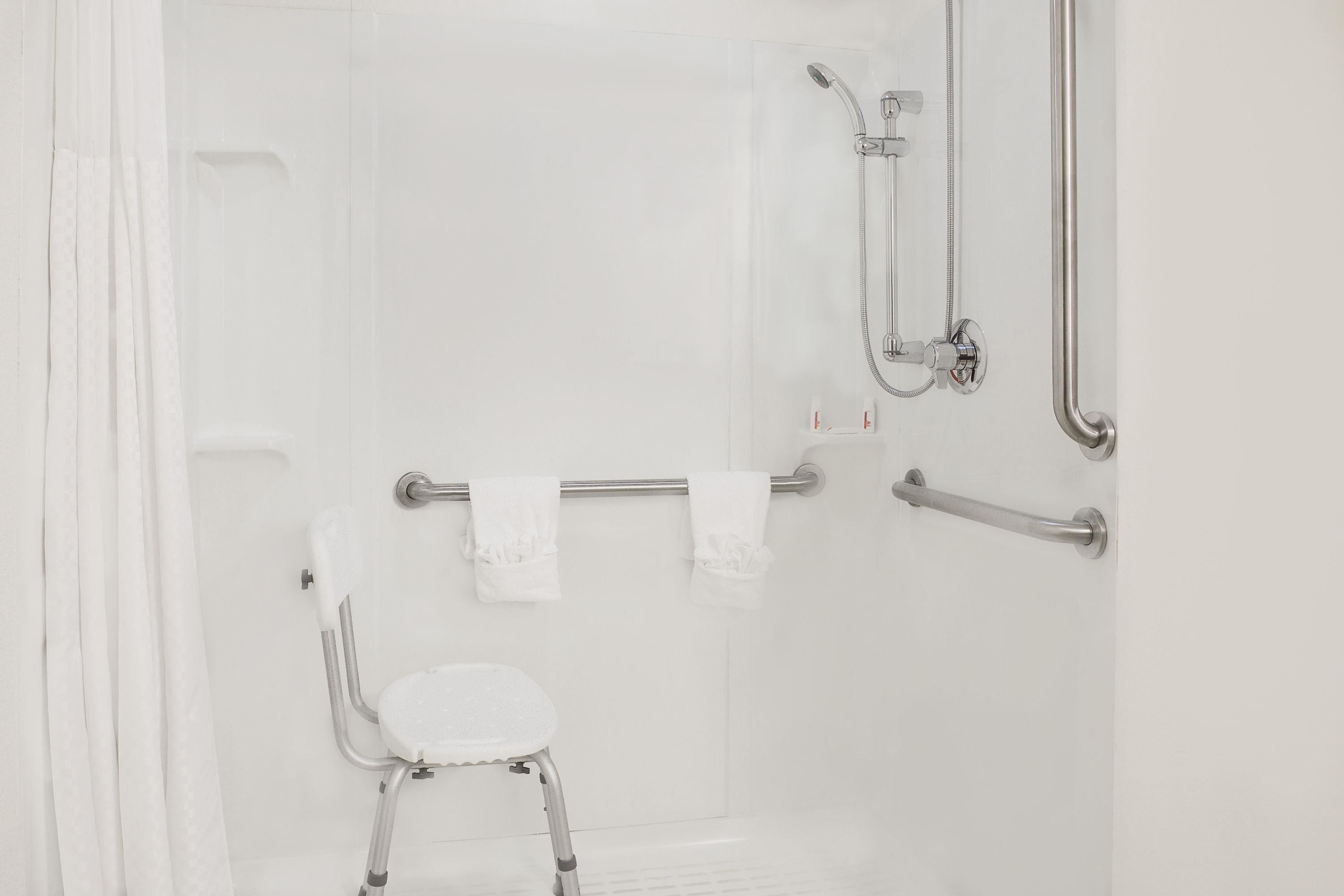 Un baño blanco con ducha, barras de apoyo y una silla blanca.