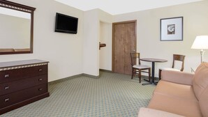 Habitación de hotel con un aparador de madera, un televisor montado en la pared, un cuadro enmarcado, una puerta, una mesita redonda con dos sillas y un sofá color beige.