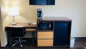 Habitación de hotel pequeña con escritorio, lámpara, microondas, refrigerador y televisor montado en la pared.