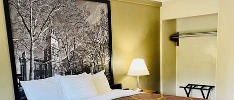 A hotel room with a bed, a nightstand with a lamp, a wall-mounted towel rack, and a large framed picture of a building.