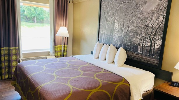 Habitación de hotel con una cama grande, una ventana con cortinas, un reloj en la pared y un cuadro con una vista urbana.