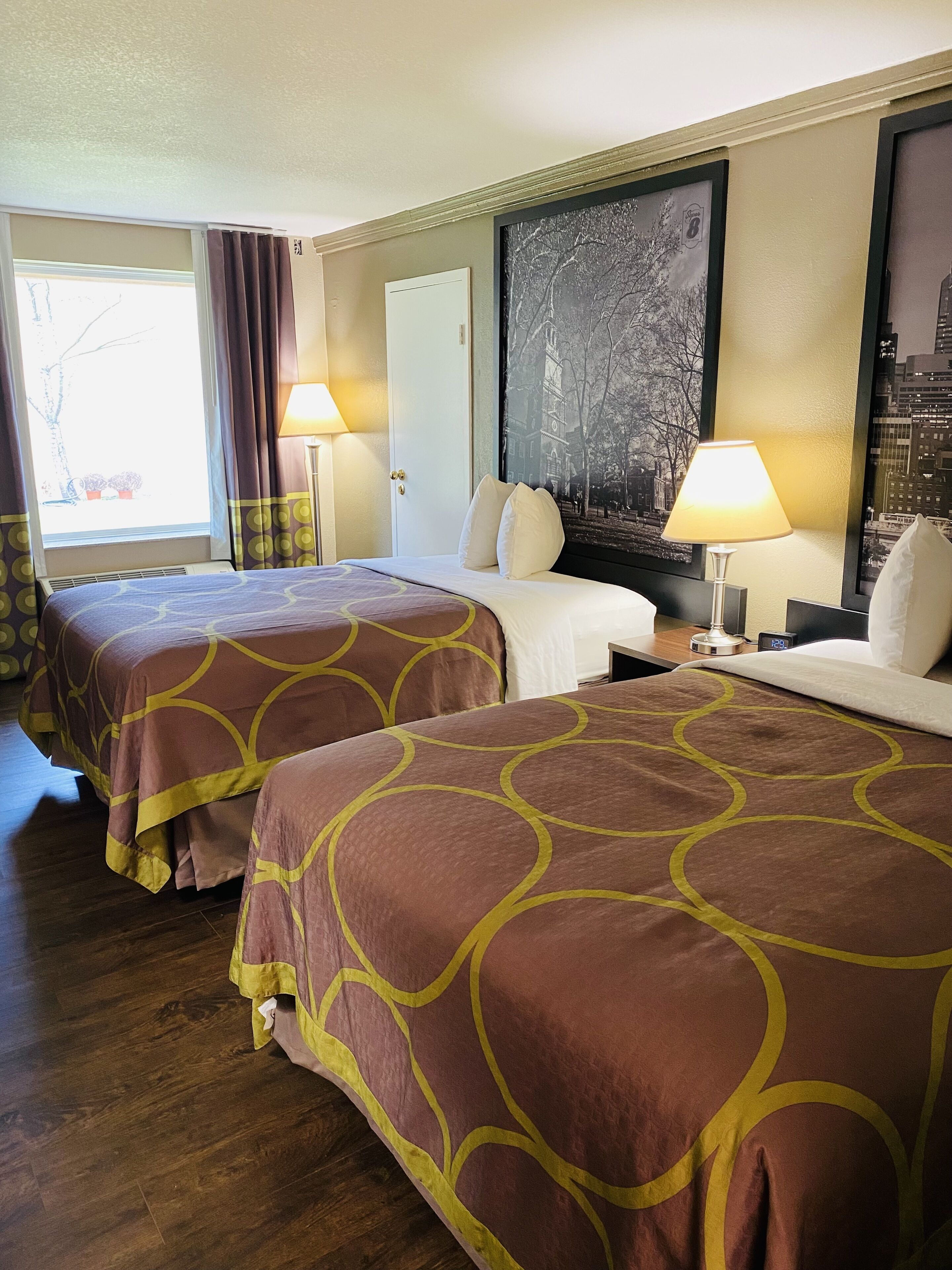 A hotel room with two beds, a large window with curtains, a wall-mounted painting, and a bedside table with a lamp.