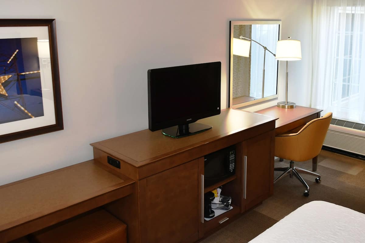 A hotel room with a desk, a television, a chair, a bed, a nightstand, a lamp, a framed picture, and a window with curtains.