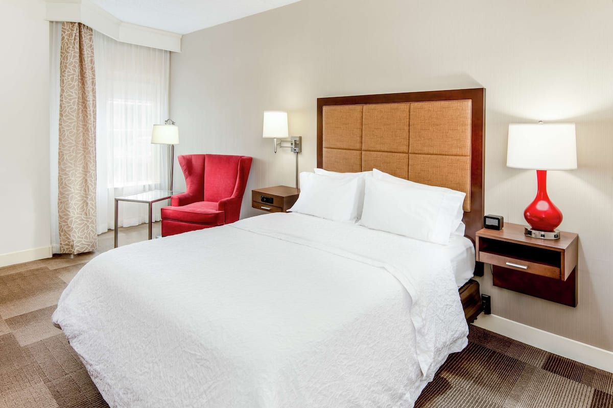 A hotel room with a large bed, a red armchair, a nightstand with a lamp, and a window with sheer curtains.
