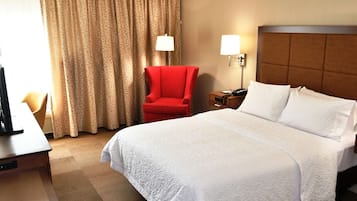 Ett hotellrum med en stor säng, ett skrivbord med en dator, en röd fåtölj, ett nattduksbord med en lampa och ett fönster med gardiner.