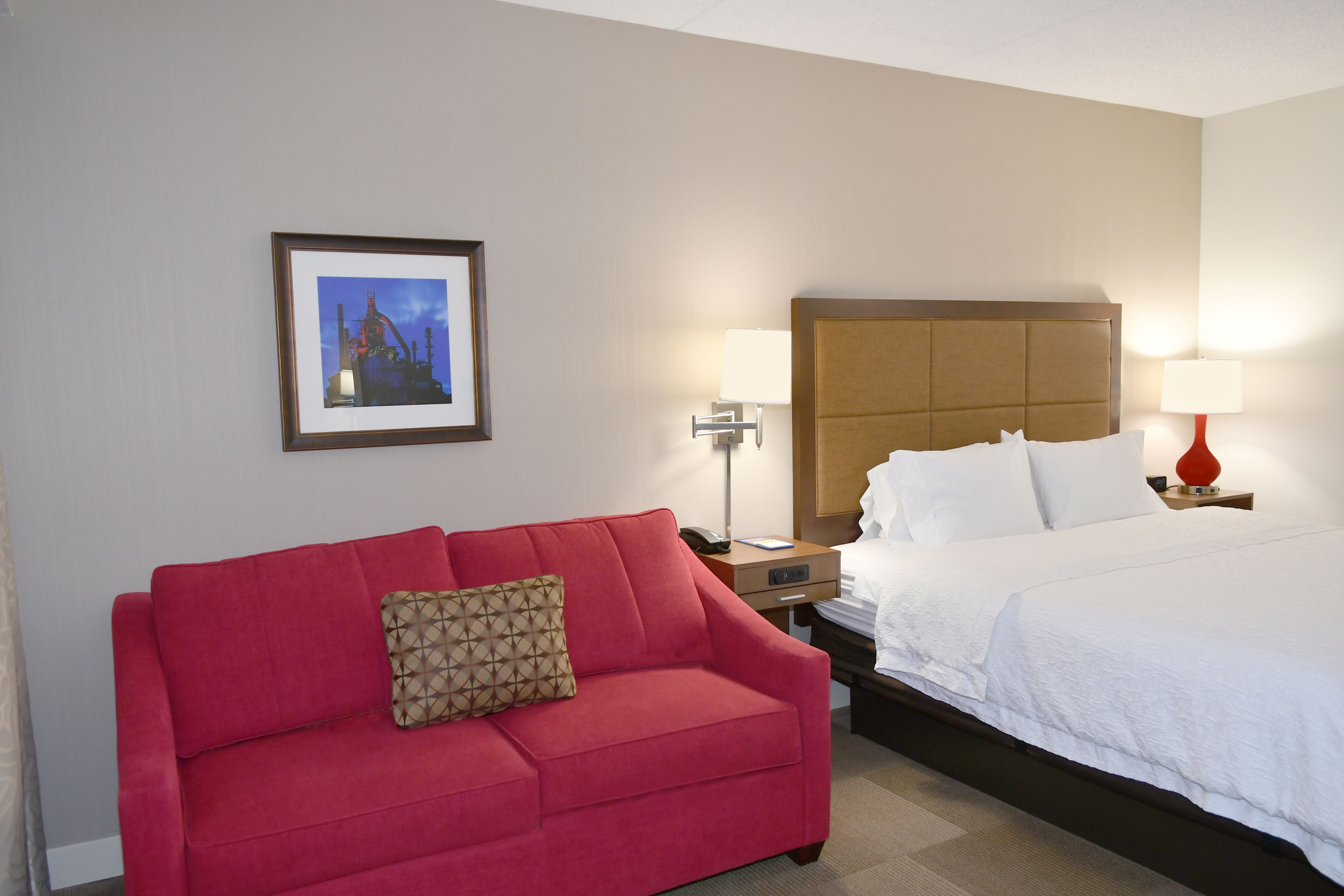 Habitación de hotel con sofá rojo, cama con ropa de cama blanca, un cuadro enmarcado en la pared y dos lámparas de noche.