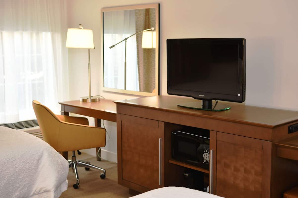 A hotel room with a bed, a desk with a computer monitor, a chair, a lamp, and a mirror.