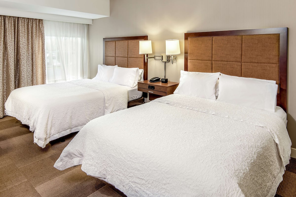 A hotel room with two beds, a nightstand with a phone and lamp, and a window with sheer curtains.