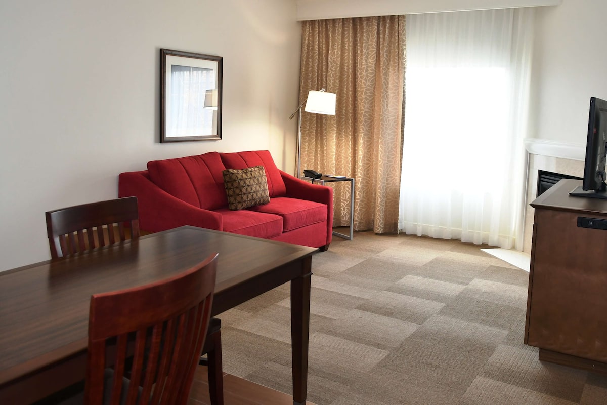 A hotel room with a red sofa, a wooden dining table with chairs, a lamp, and a television on a cabinet.