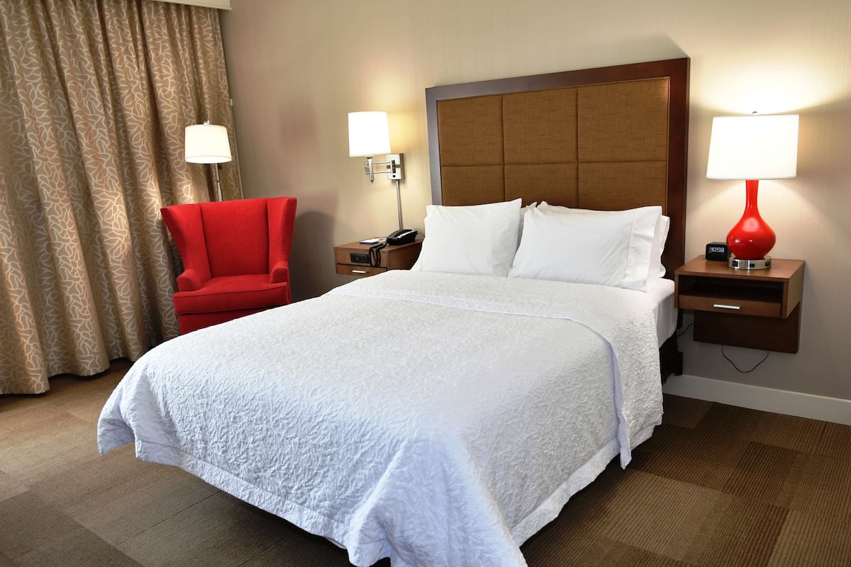 A hotel room with a large bed, a red armchair, two bedside tables with lamps, and a wooden headboard.