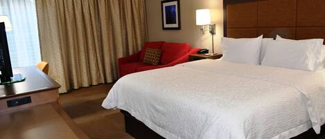 A hotel room with a large bed, a desk with a lamp, a red chair, and a window with curtains.