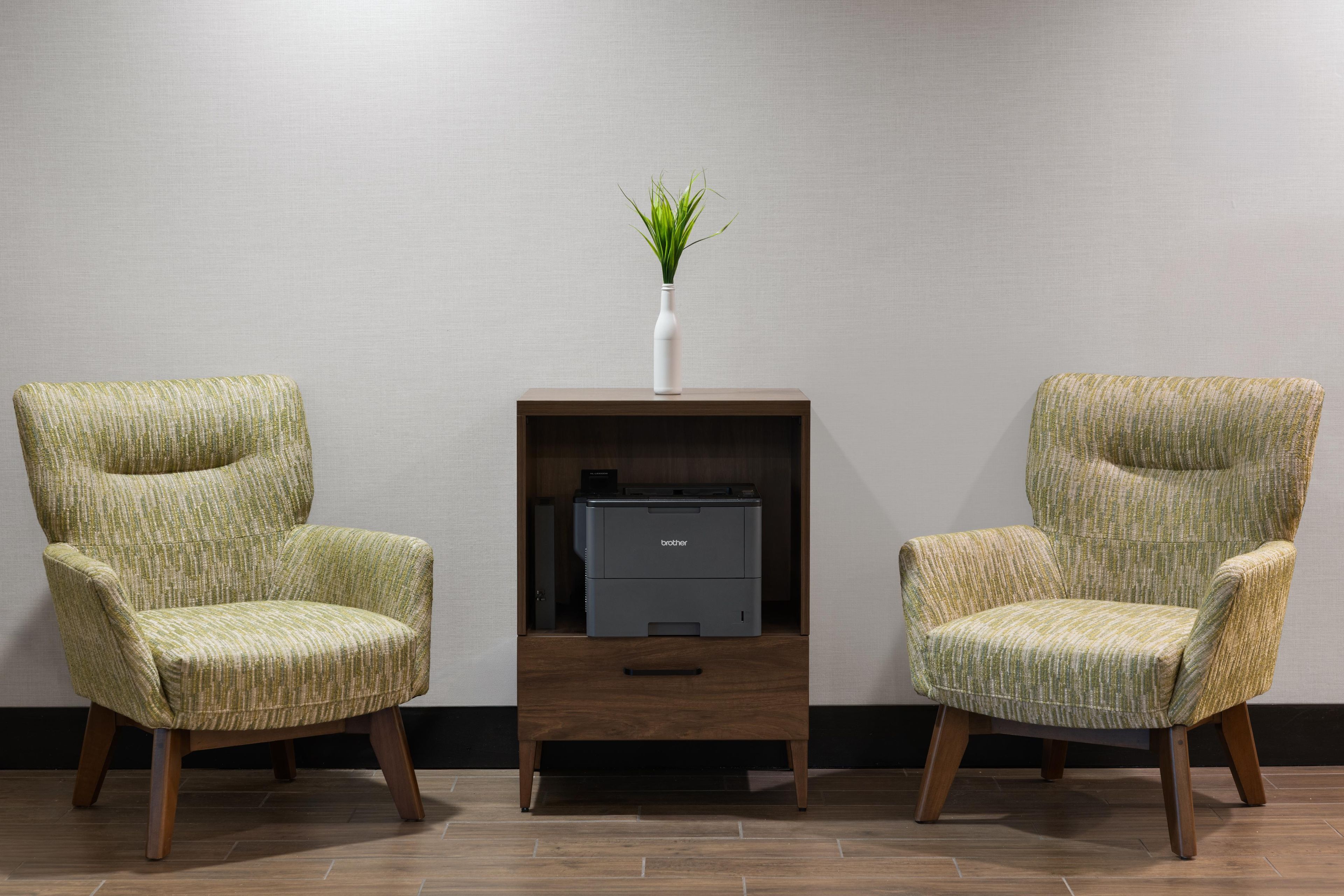 Two armchairs with a patterned upholstery, a small wooden side table with a printer, and a white vase with green foliage.