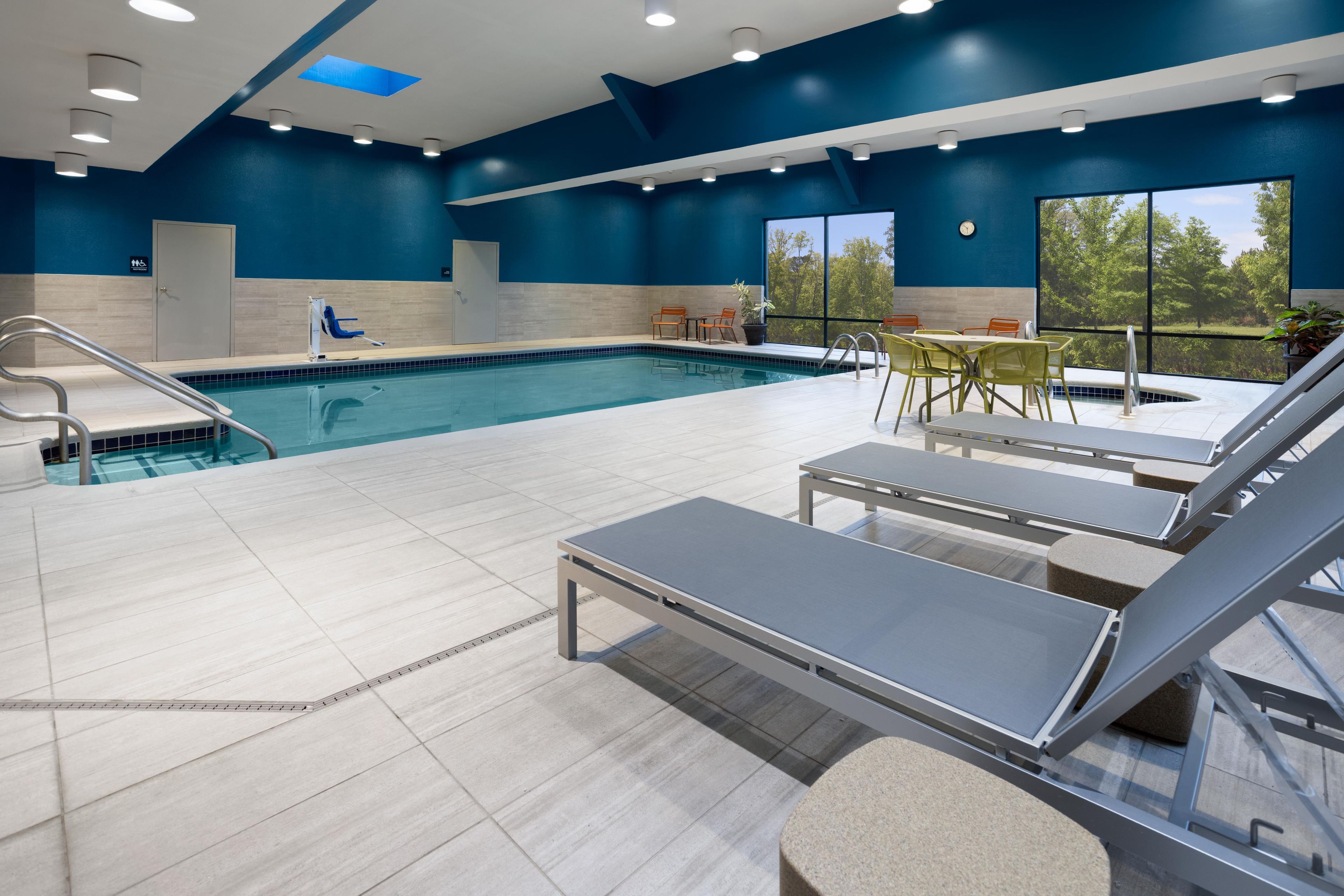 Piscina interior moderna con vistas despejadas al exterior, zona de estar con mesas y silla de salvavidas.