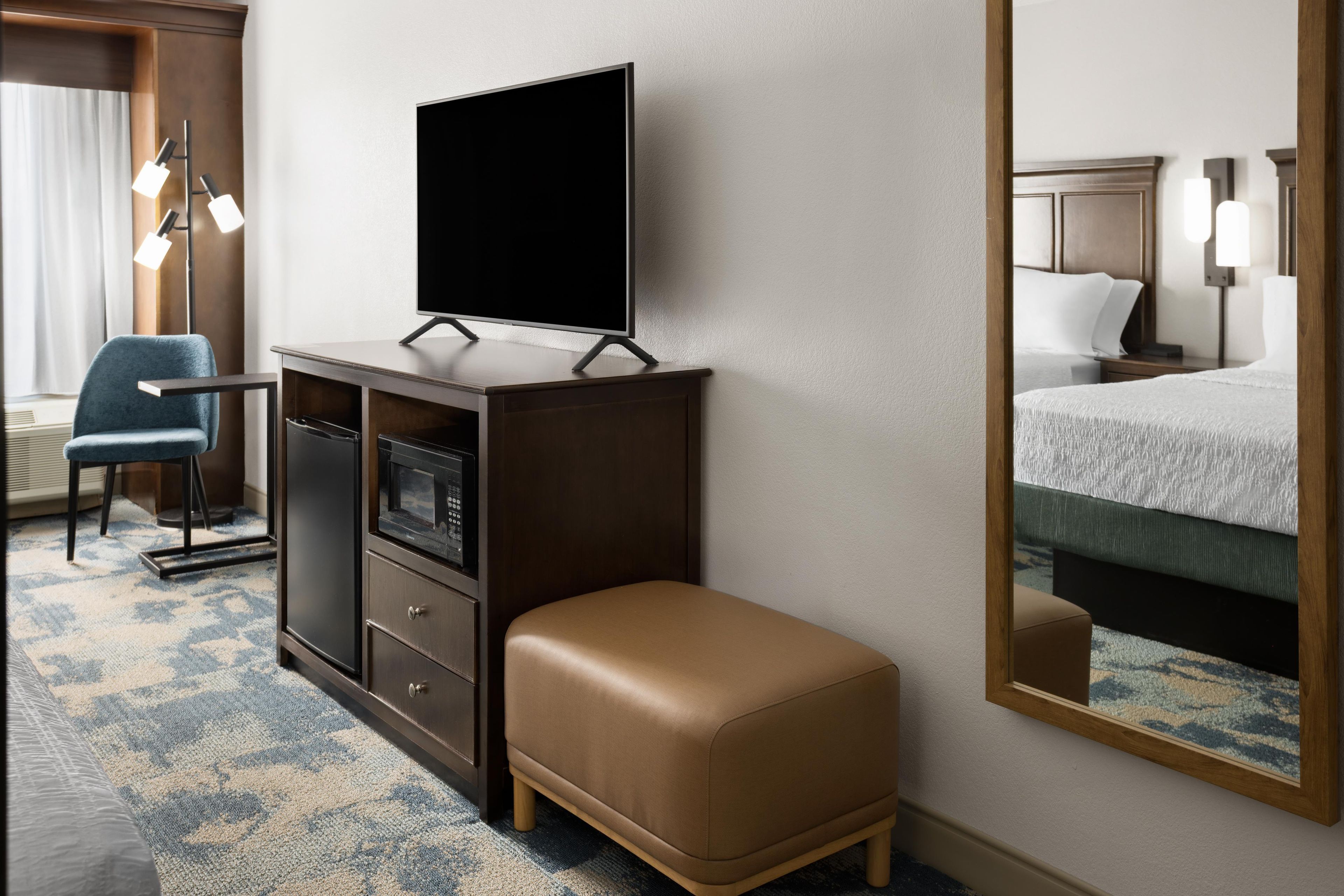 Habitación de hotel con televisor de pantalla plana sobre un mueble de madera, un sillón azul, un otoman marrón, un espejo grande que refleja la cama y una mesita de noche, además de una alfombra estampada.