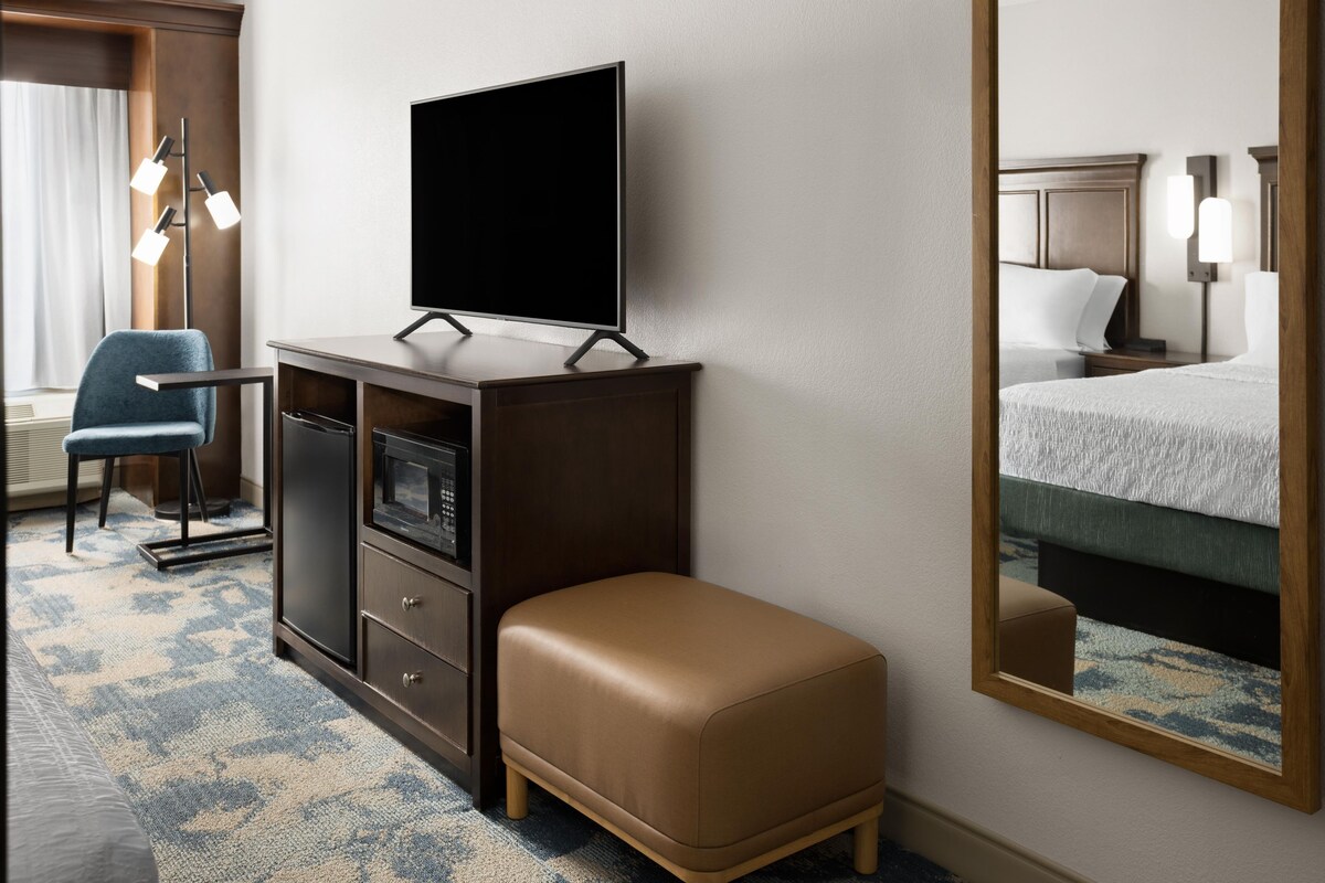 A hotel room with a flat-screen TV on a wooden cabinet, a blue armchair, a brown ottoman, a large mirror reflecting the bed and a nightstand, and a patterned carpet.