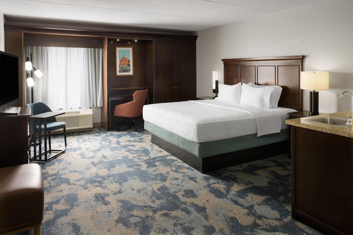 A hotel room with a bed, desk, chair, and a small kitchenette.