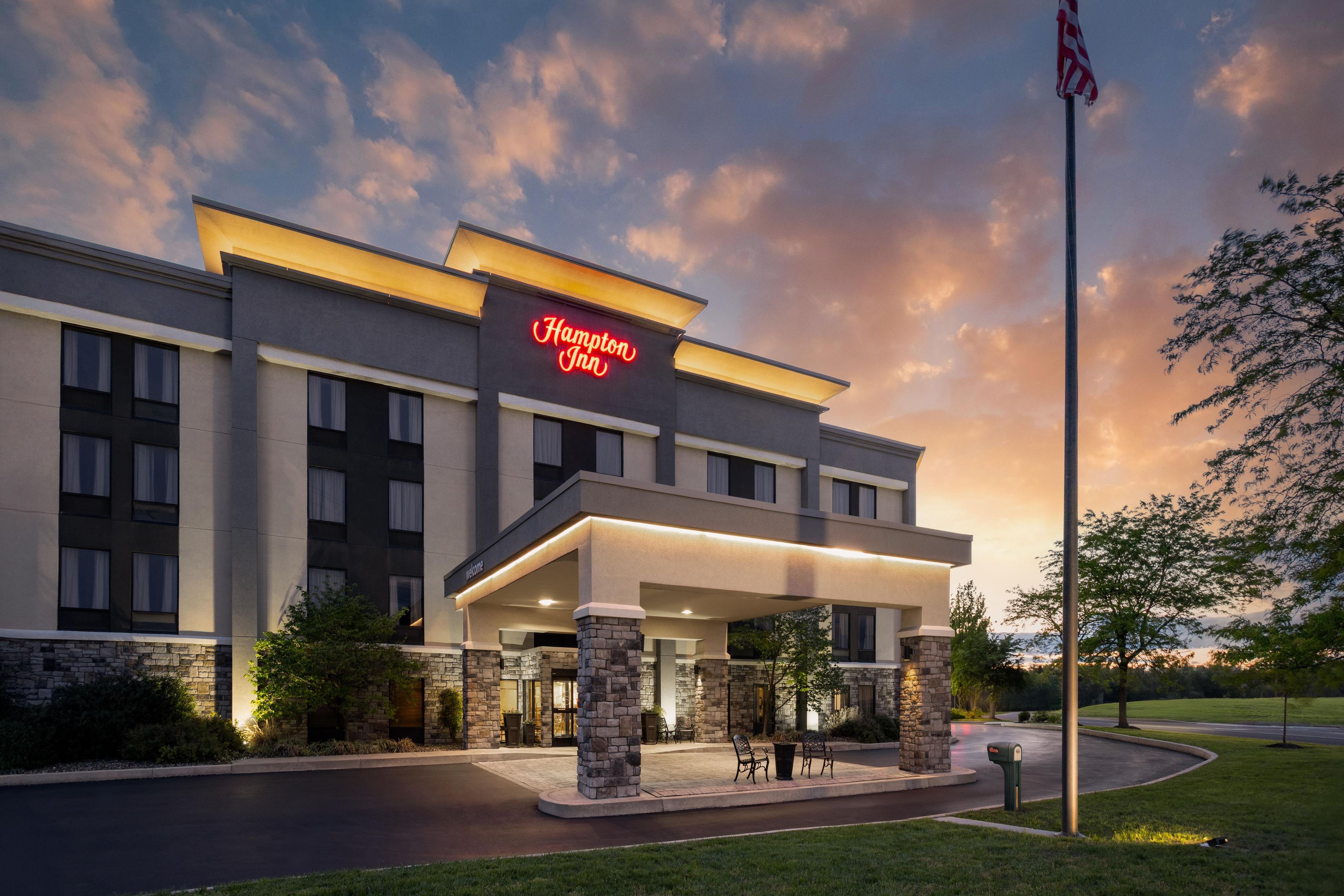 A Hampton Inn hotel with a modern design, featuring a prominent neon sign, a covered entrance, and a flagpole.