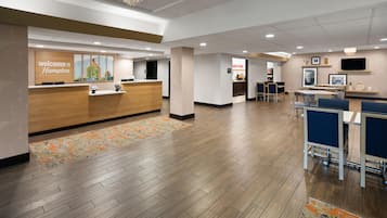 A hotel reception area with a wooden reception desk, a 'Welcome to Hampton' sign, and a view into the dining area with tables and chairs.