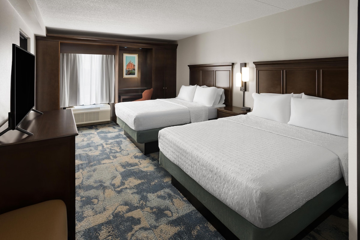 A hotel room with two beds, a desk, a TV, and a painting on the wall.
