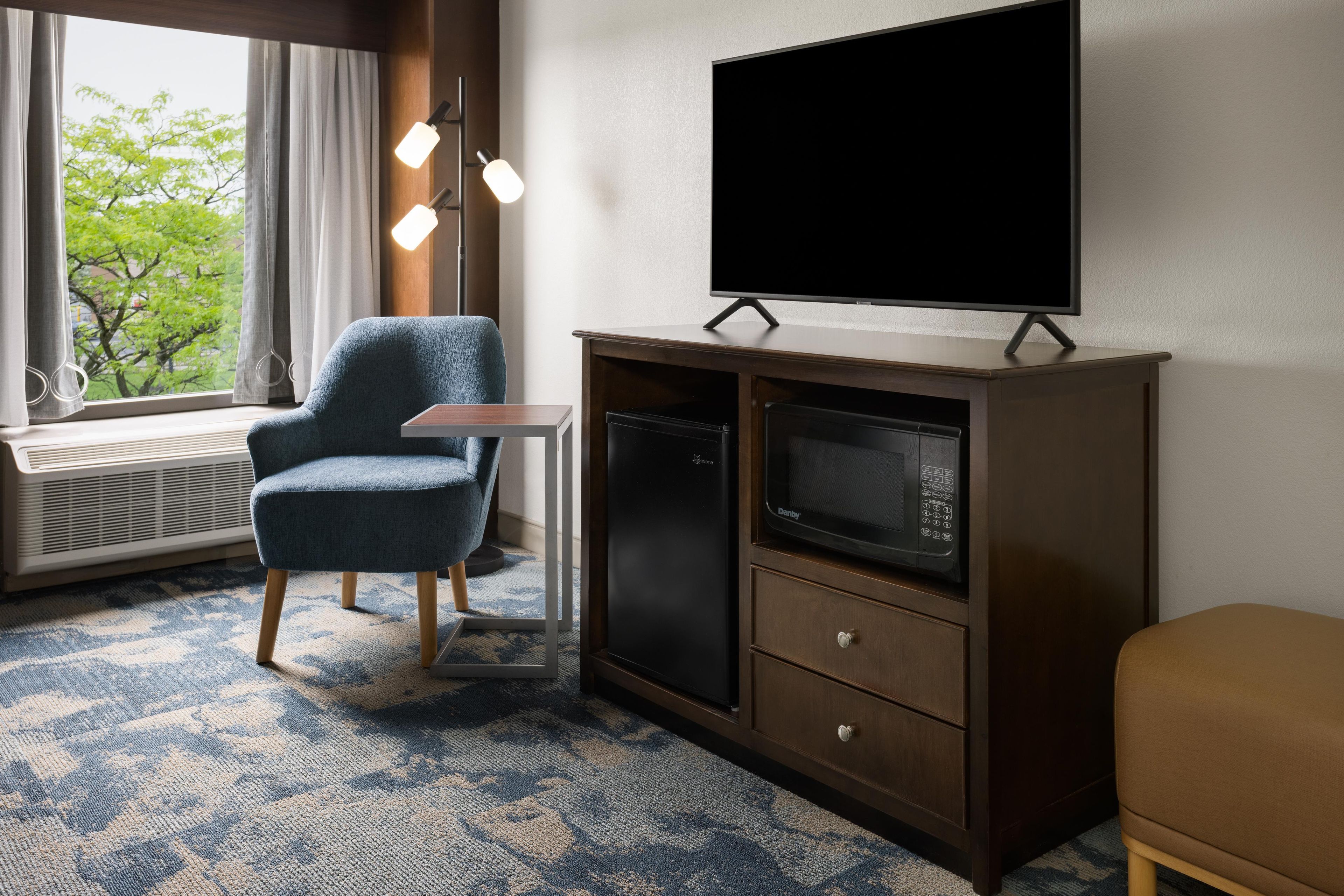 Ein Hotelzimmer mit Flachbildfernseher auf einer Holzkabine, einem blauen Sessel, einem kleinen Tisch und einem Fenster mit Vorhängen.