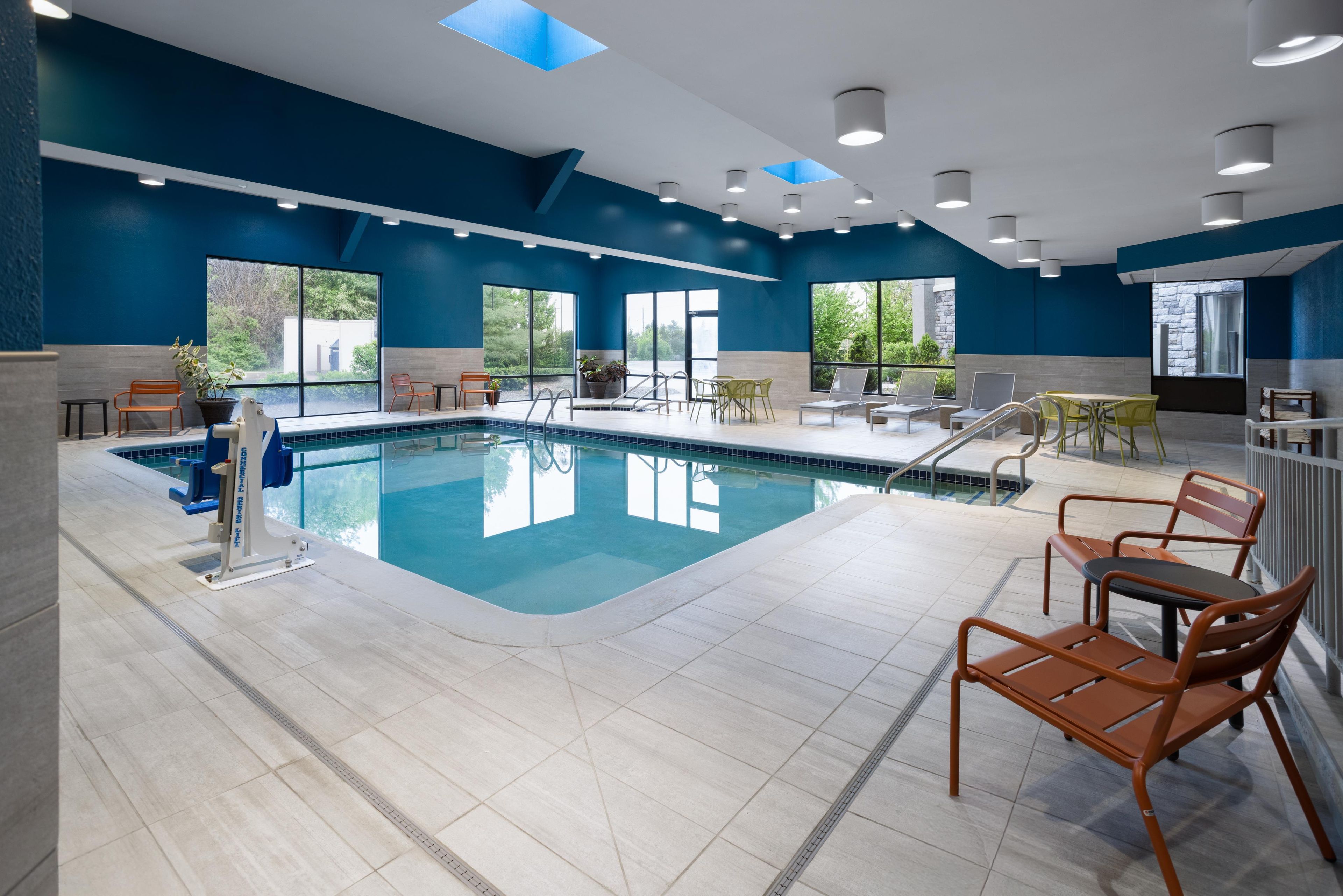 Una piscina interior moderna con agua azul cristalina, rodeada de sillones y mesas, y amplias ventanas que ofrecen vistas a la vegetación.