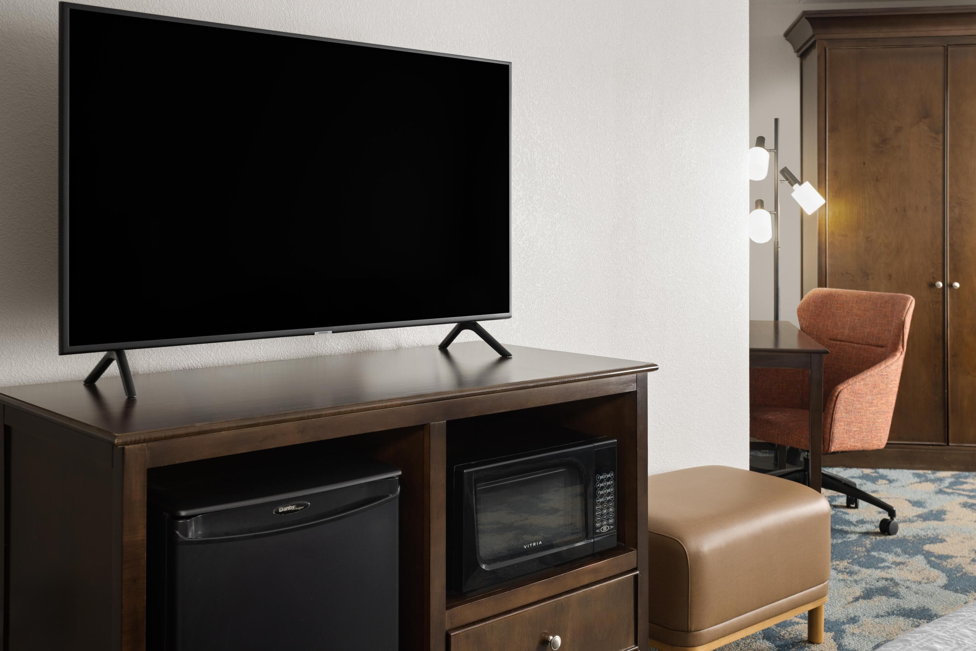 A hotel room with a flat-screen TV mounted on a wooden cabinet, a black microwave, a brown leather ottoman, and a wooden wardrobe with a mirror and lights.