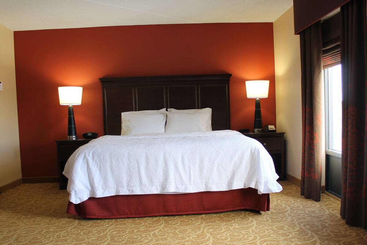 Una habitación de hotel con una cama grande, dos lámparas de noche y un ventanal con cortinas.