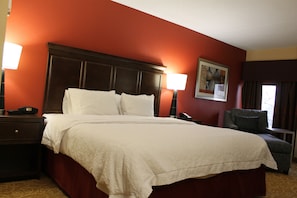 A hotel room with a large bed, a nightstand, a lamp, a chair, and a painting on the wall. - Hampton Inn Gettysburg (Gettysburg)