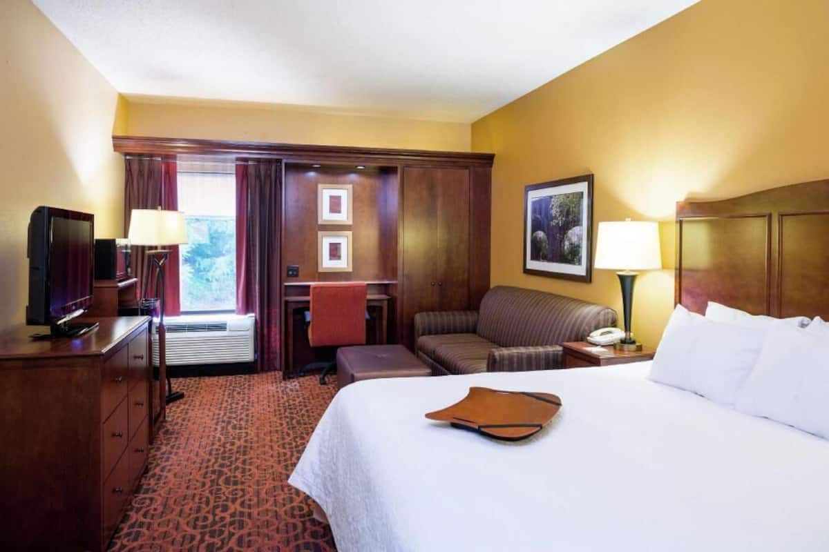 Habitación de hotel con cama, escritorio, silla, televisor, lámpara y ventana con cortinas.