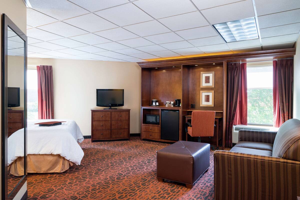 Habitación de hotel con cama, televisor, kitchenette pequeña, sofá y ventana grande con cortinas.