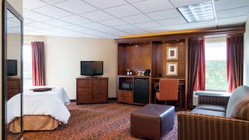 A hotel room with a bed, a television, a small kitchenette, a sofa, and a large window with curtains.