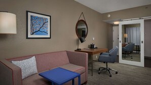 Habitación de hotel con sofá, una mesita, escritorio con silla, lámpara, cuadro enmarcado, espejo y puerta que da a otra habitación.