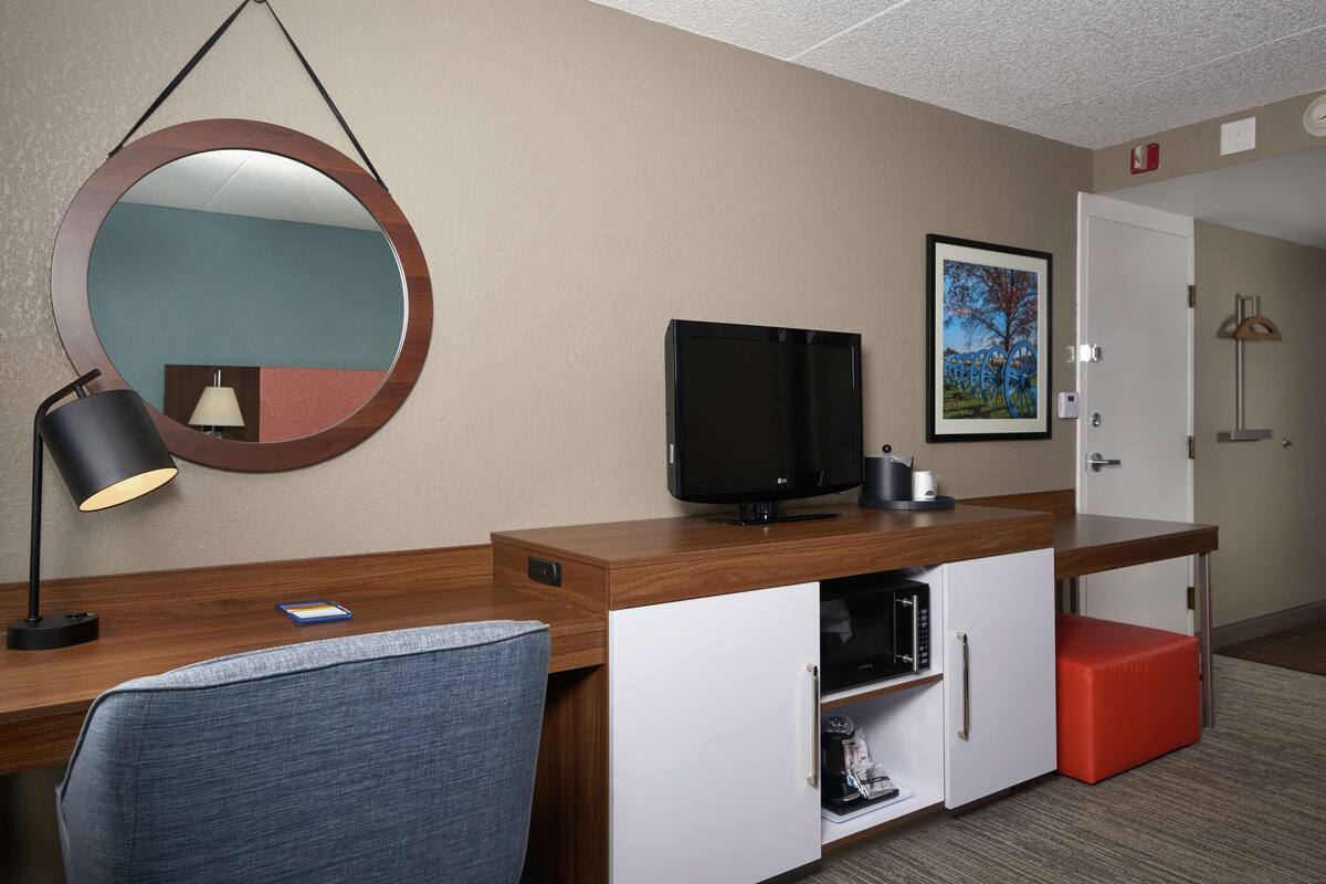 Una habitación de hotel con un escritorio de madera, un televisor, un espejo, un reposapiés rojo, un armario blanco y un cuadro en la pared.