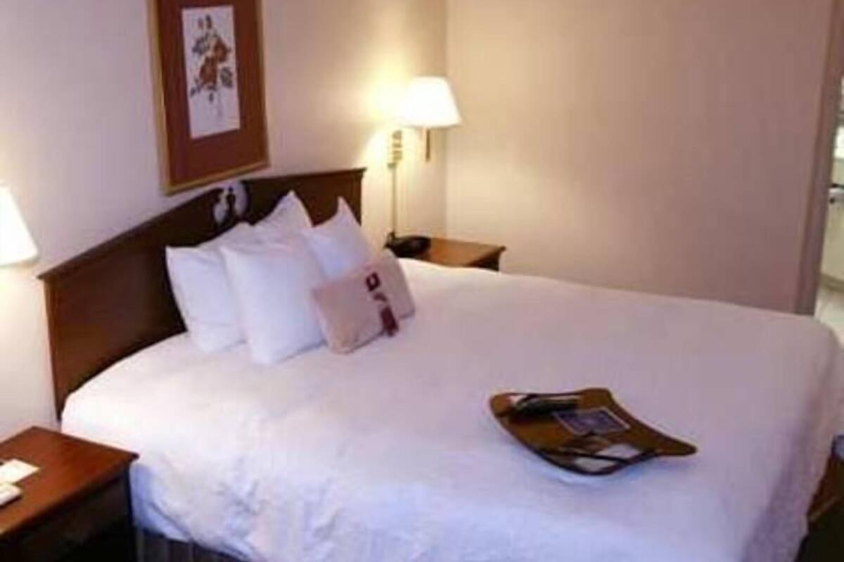 Una cama bien tendida con ropa blanca, cabecera de madera, mesita de noche con lámpara y un cuadro enmarcado en la pared.