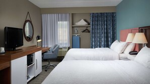 Habitación de hotel con dos camas, un escritorio con televisor, una silla, armario, espejo, ventana con cortinas y mesita de noche con lámparas.