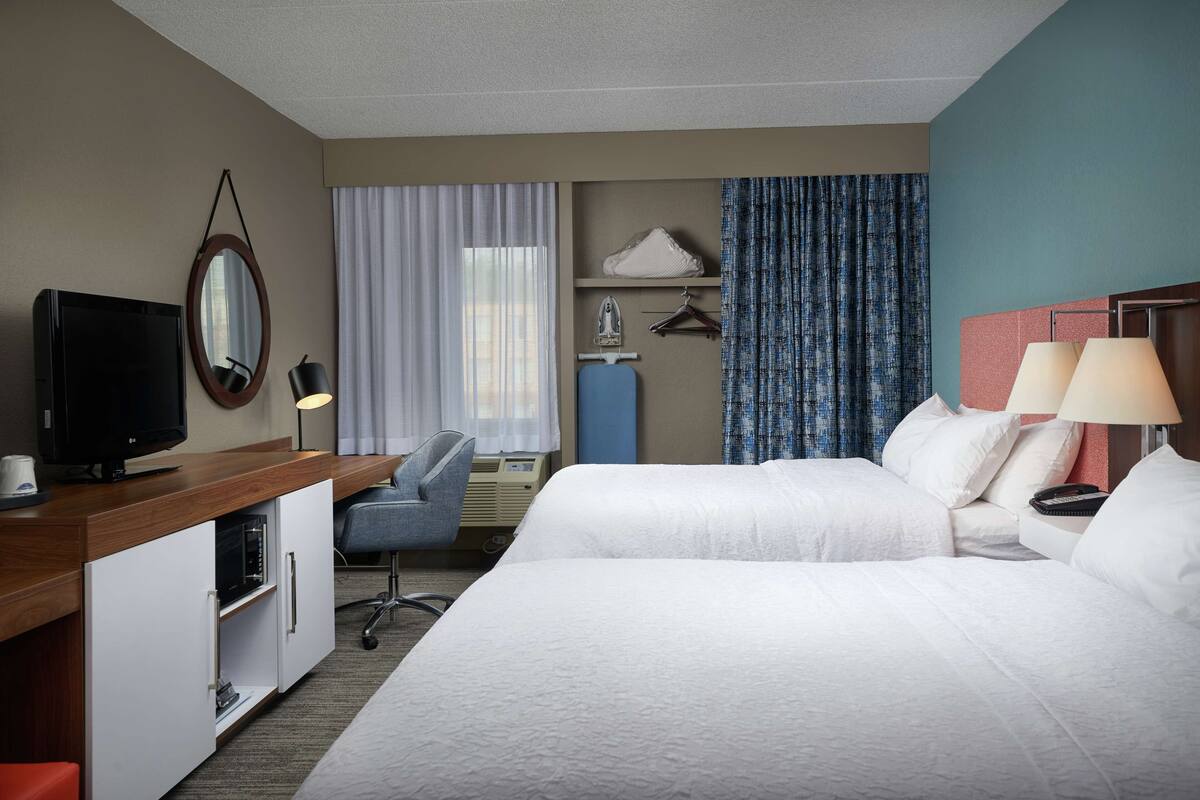 Habitación de hotel con dos camas, un escritorio con televisor, una silla, armario, espejo, ventana con cortinas y mesita de noche con lámparas.