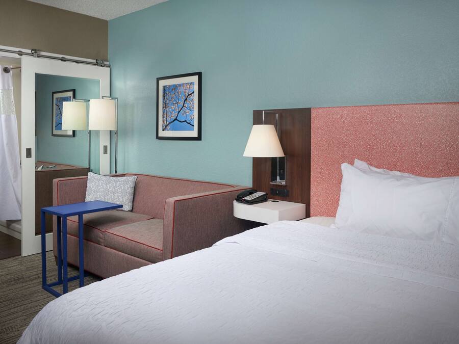 A hotel room with a bed, a desk, a chair, a nightstand, a lamp, and a picture on the wall.