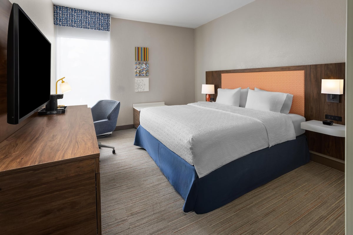 A hotel room with a large bed, a desk with a TV, a chair, and a nightstand with a lamp.